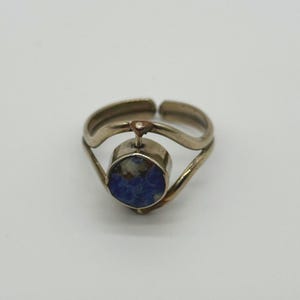 May include: A silver-toned ring with a round, deep blue stone set in the center. The ring has a unique design with a curved band and adjustable sizing. The stone is the focal point, set against a simple, elegant metalwork.