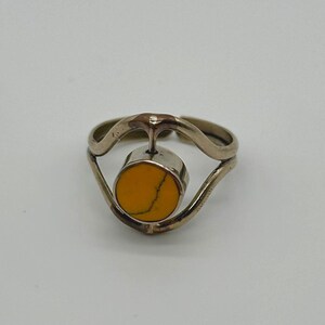 May include: A silver ring with a round, yellow-orange stone set in the center. The ring has a unique, open design with curved lines. The stone is a bright, solid color, and the metal has a polished finish. The ring is a piece of jewelry.