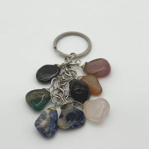 May include: A keychain with a silver ring and chain holding a cluster of polished gemstones. The stones are various colors, including black, green, blue, pink, and brown. The gemstones are smooth and rounded.