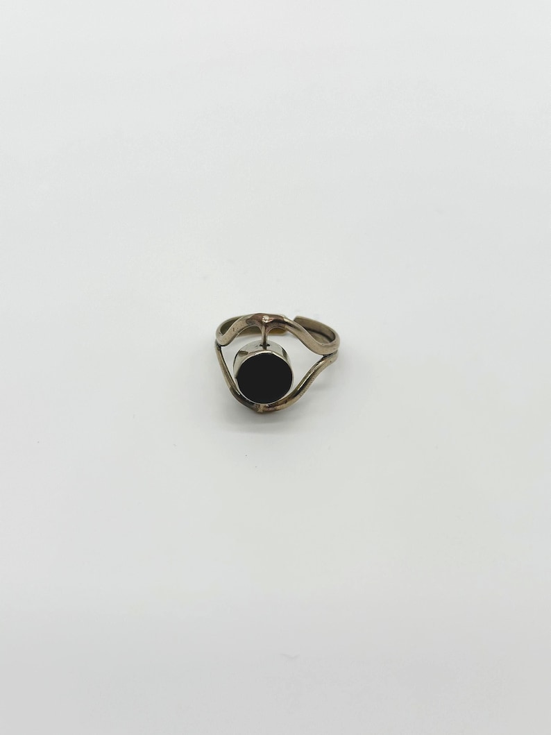 May include: A silver ring with a black circular stone set in the center. The ring has a unique, wavy design on the band. The ring is displayed against a white background.