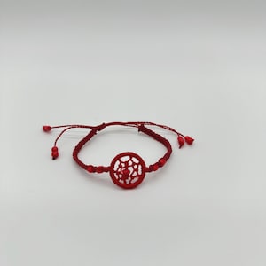 May include: A red macrame bracelet with a circular, openwork design in the center. The bracelet features small red beads and adjustable ties. The bracelet is displayed on a white background.