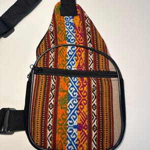 May include: A colorful sling bag with a woven fabric design. The bag features a black strap, zipper, and trim. The fabric has vertical stripes in red, orange, yellow, blue, and white. The bag is designed to be worn across the body.