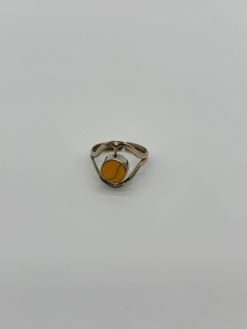 May include: A silver ring with a round, yellow-orange stone set in the center. The ring has a unique, open design with curved lines. The stone is a bright, solid color, and the metal has a polished finish. The ring is a piece of jewelry.