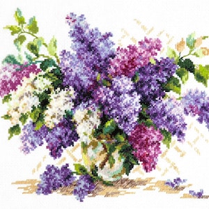 May include: Cross-stitch embroidery of a lilac bouquet in a vase. The flowers are in shades of purple, white, and pink, with green leaves. The vase is clear with a floral design, and the background is white.