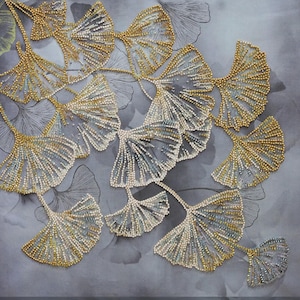 Peut inclure: Gros plan sur une œuvre d'art en perles représentant une branche de feuilles de ginkgo. Les feuilles sont faites de petites perles irisées dans des tons d'or et d'argent, créant un effet chatoyant. L'arrière-plan est d'un gris doux.