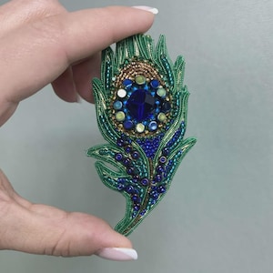 Op de afbeelding: Handgemaakte pauwenveerbroche met ingewikkeld kralenwerk. De broche heeft een grote, facetgeslepen blauwe edelsteen in het midden, omringd door kleinere blauwe, groene en gouden kralen. De omtrek van de veer is afgebakend met groene kralen.