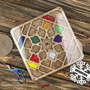 Può includere: Un organizer per artigianato in legno con vari scomparti pieni di perline colorate. L'organizer ha un design geometrico e un coperchio trasparente. Sono visibili anche forbici iridescenti e un ritaglio a forma di fiocco di neve.