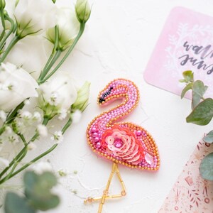 Peut inclure: Une broche en forme de flamant rose en perles avec des pattes en fil d'or et des paillettes roses. La broche est sur une surface blanche avec des fleurs blanches et une carte rose avec le mot "walking" écrit dessus.