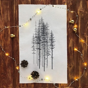 Könnte beinhalten: Eine Kreuzstich-Stickerei einer Waldszene mit hohen, schlanken Bäumen in Grau- und Schwarztönen. Das Kunstwerk befindet sich auf einem weißen Stoff, mit zwei Tannenzapfen und Lichterketten mit goldenen Glühbirnen und silbernen Perlen auf einem Holzgrund.