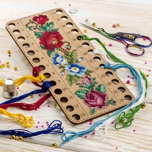 Puede incluir: Un organizador de hilos de madera con 26 agujeros para guardar el hilo de bordar. El organizador tiene un diseño floral con flores rojas, azules y verdes. Hay varios hilos de diferentes colores, incluyendo rojo, azul, amarillo y verde, envueltos alrededor del organizador.