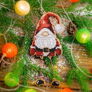 May include: A red and white Santa Claus bauble with a white beard and a black belt. The bauble is hanging from a Christmas tree with green branches and white lights.
