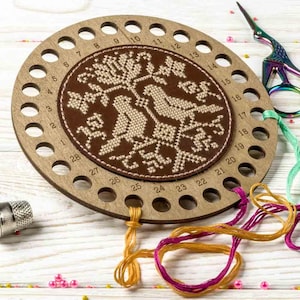 Może przedstawiać: Drewniany organizer na nici do haftu ze wzorem wyszywanego ptaka. Okrągły organizer ma ponumerowane otwory do przechowywania nici i jest otoczony kolorowymi nićmi do haftu i parą nożyczek.