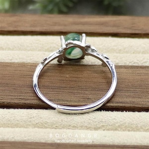 May include: A silver ring with a green gemstone set in a delicate floral design. The ring is on a wooden surface.
