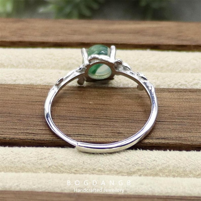 May include: A silver ring with a green gemstone set in a delicate floral design. The ring is on a wooden surface.
