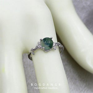May include: A silver ring with a green agate stone set in a delicate branch design. The ring is on a white mannequin hand.