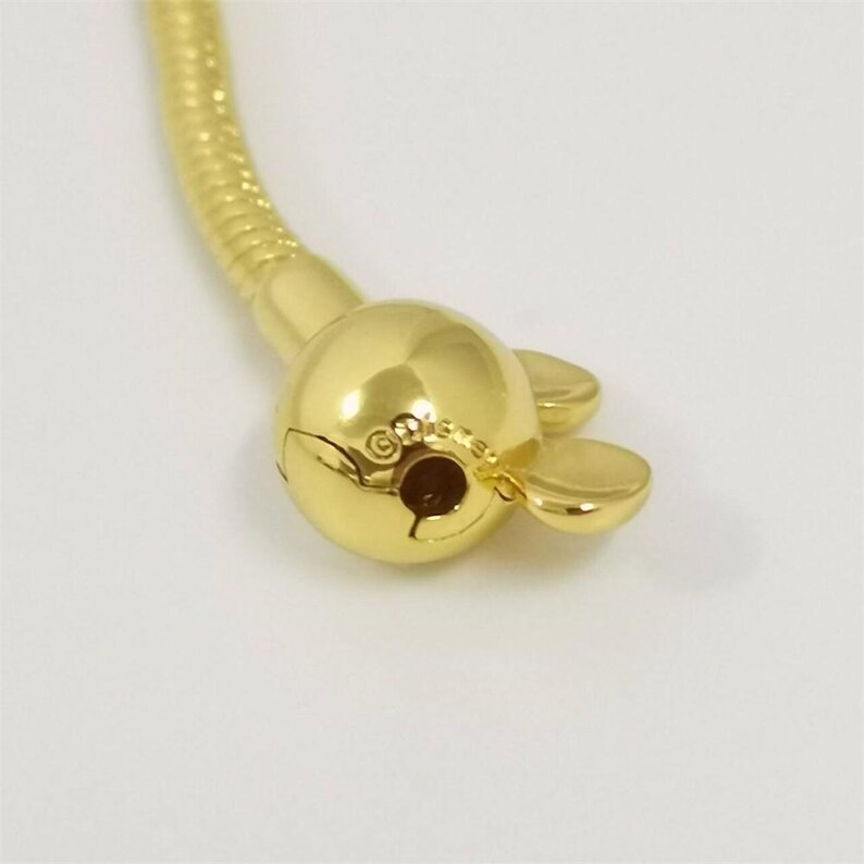 Op de afbeelding: Close-up van een goudkleurige armbandsluiting. De sluiting is rond met twee kleine, afgeronde verlengingen aan weerszijden. De armbandketting is een flexibele, goudkleurige slangenketting.