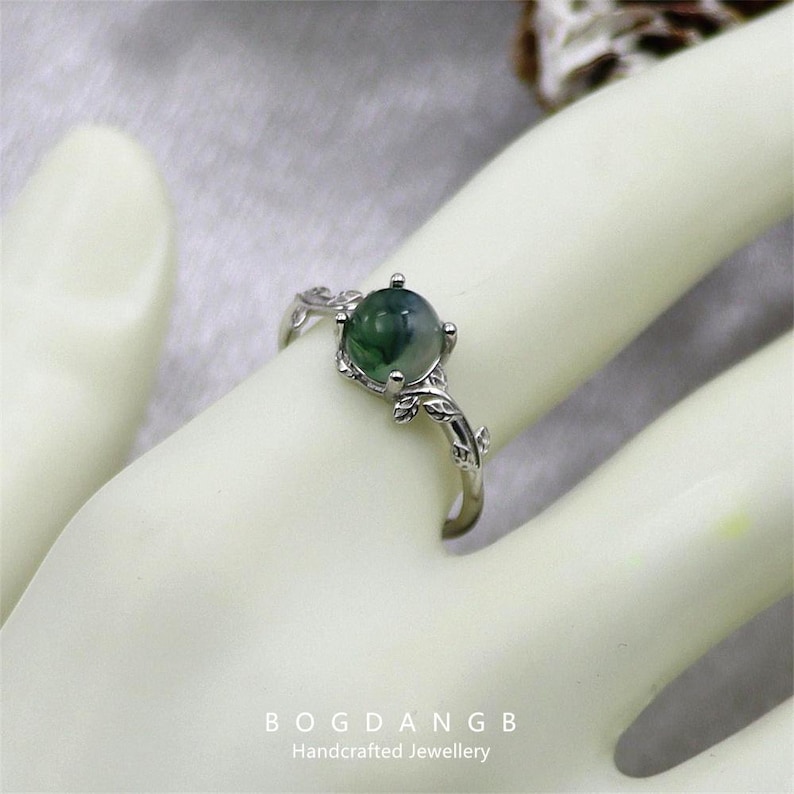 May include: A silver ring with a green agate gemstone and a leafy design. The ring is on a mannequin hand.