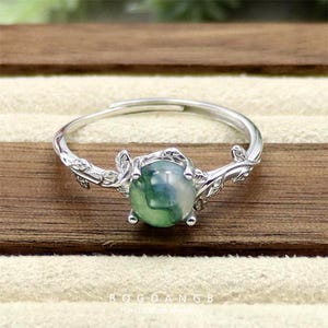 May include: A silver ring with a green agate stone set in a delicate branch design. The ring features a leafy branch design with a round green agate stone in the center.