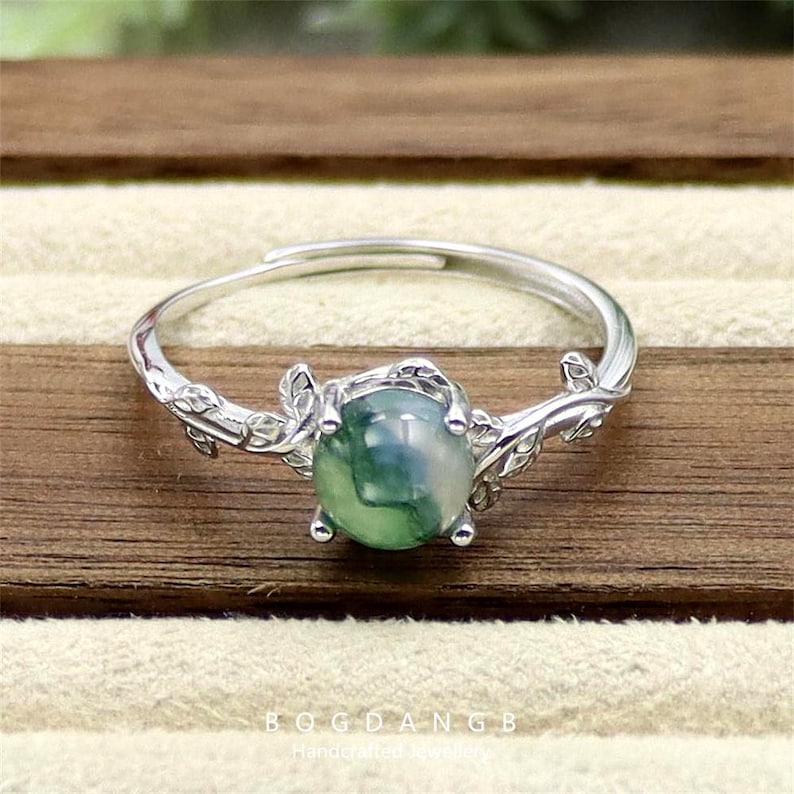 May include: A silver ring with a green agate stone set in a delicate branch design. The ring features a leafy branch design with a round green agate stone in the center.