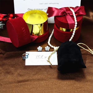 May include: A red velvet jewelry box with a gold lid, a pearl necklace, a pair of flower-shaped earrings, a ring with a blue stone, and a small black velvet pouch. The box has a white ribbon and a tag that says "cool and refreshing" and "One summer of".