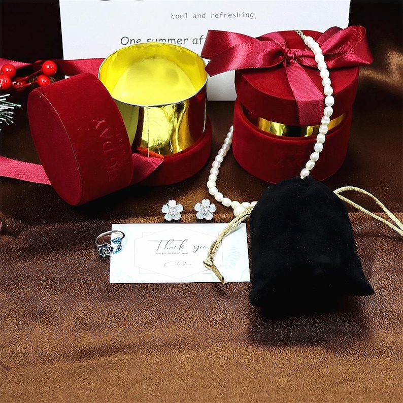 May include: A red velvet jewelry box with a gold lid, a pearl necklace, a pair of flower-shaped earrings, a ring with a blue stone, and a small black velvet pouch. The box has a white ribbon and a tag that says "cool and refreshing" and "One summer of".
