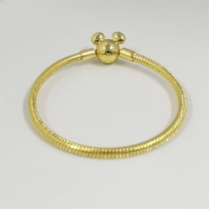 Op de afbeelding: Een goudkleurige armband met een Mickey Mouse-vormige sluiting. De armband heeft een textuur, slangachtig ontwerp. De sluiting heeft een rond midden met twee kleine ronde oren bovenop. De armband staat op een witte achtergrond.