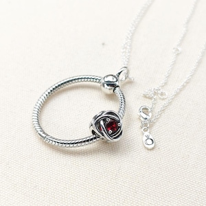 May include: A silver necklace featuring a charm with a red gemstone rose design. The necklace has a circular pendant with a textured pattern and a smaller chain with a clasp and a charm.