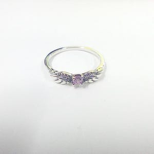 May include: A silver ring with a central, oval-cut, pale purple gemstone. The ring has wing-shaped details either side of the stone, giving it an angelic appearance. The ring is displayed on a white surface.