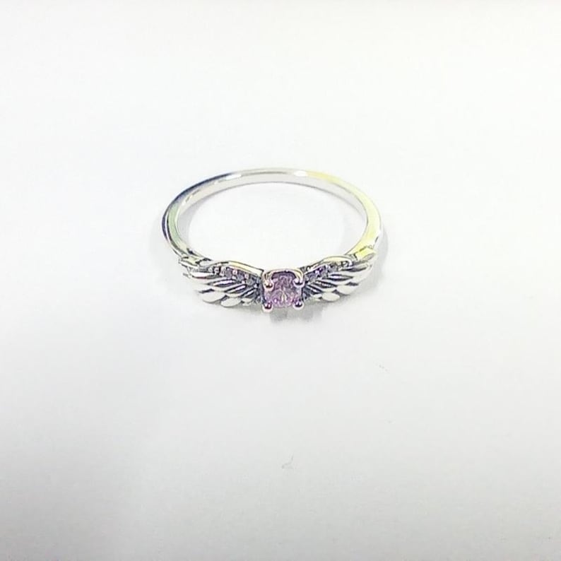 May include: A silver ring with a central, oval-cut, pale purple gemstone. The ring has wing-shaped details either side of the stone, giving it an angelic appearance. The ring is displayed on a white surface.