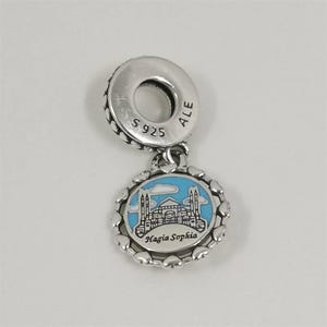 Può includere: Un charm in argento con un ciondolo rotondo raffigurante Santa Sofia a Istanbul, Turchia, su uno sfondo di cielo blu. Il charm è circondato da un bordo a forma di cuore e presenta la scritta "Hagia Sophia" sul ciondolo.