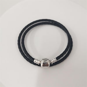 May include: Double-wrap black braided leather bracelet with a silver clasp. The clasp is engraved with "PAN". The bracelet is displayed on a white surface. The bracelet is approximately 40 cm in length.