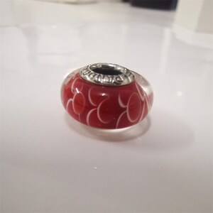 May include: A red and clear glass bead with white circular patterns. The bead features a silver-coloured metal cap. This decorative bead is likely for a bracelet or necklace.