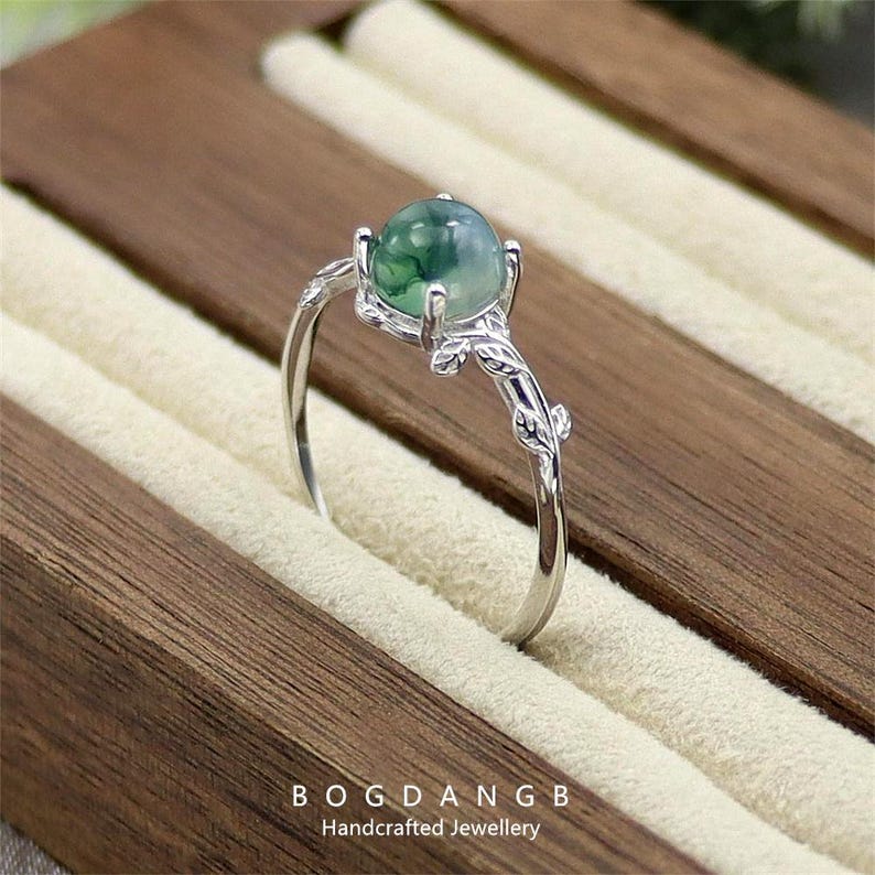 May include: A silver ring with a green agate gemstone and a delicate branch design. The ring is displayed in a wooden ring box.