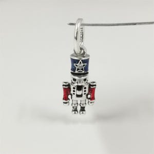 May include: A silver charm in the shape of a nutcracker soldier. The charm has a blue and silver hat with a star and a clear gem. The arms are red, and the charm is suspended from a silver loop.
