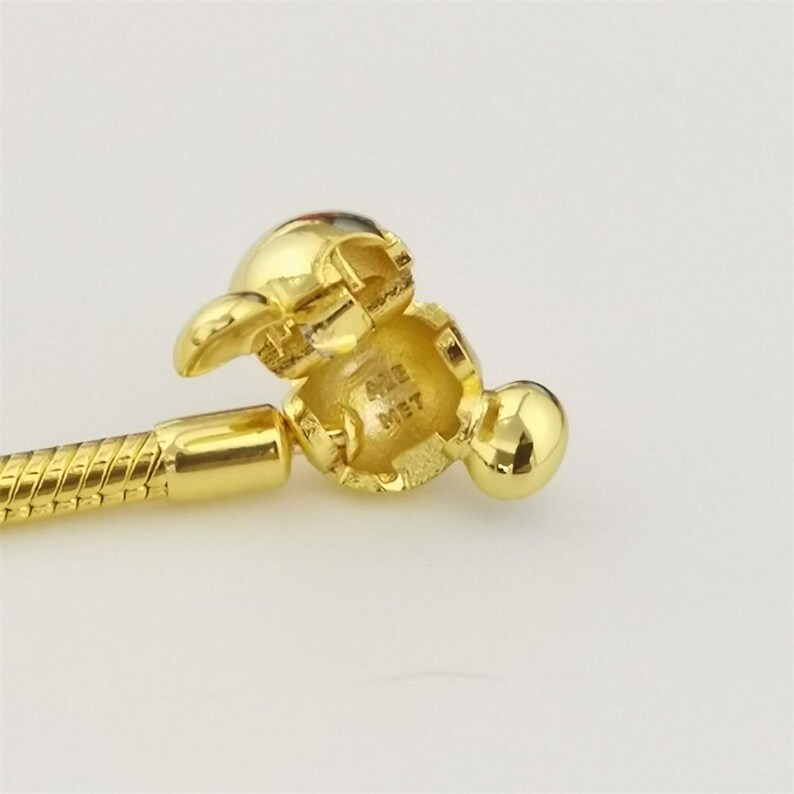 Op de afbeelding: Een goudkleurige bedelarmbandsluiting, met een gedetailleerd ontwerp en een scharnierend openingsmechanisme. De sluiting is verbonden met een gouden slangenkettingarmband. De tekst "ALE MET" is zichtbaar op de sluiting.