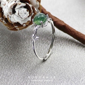 May include: A silver ring with a green agate stone and a delicate leaf design. The ring is set on a white and brown background.