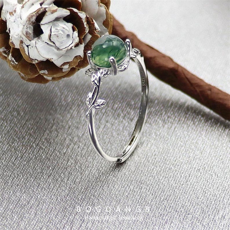 May include: A silver ring with a green agate stone and a delicate leaf design. The ring is set on a white and brown background.