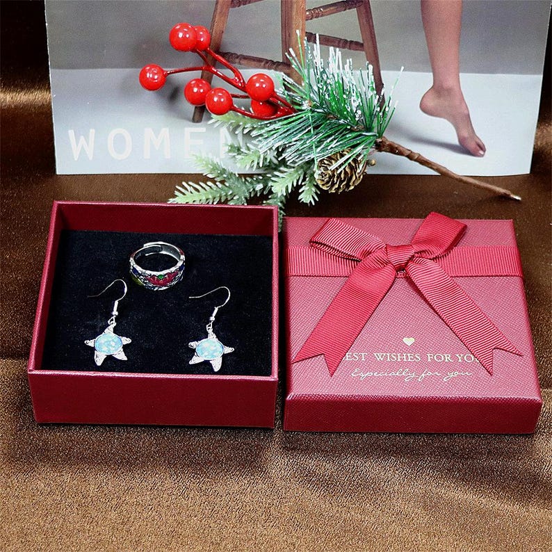 May include: A red gift box with a red bow and the text "Best Wishes For You Especially For You". The box contains a silver ring and a pair of silver earrings with blue and green stones.
