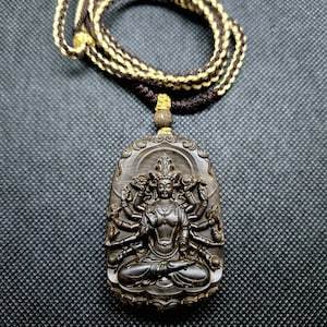 May include: A dark brown wooden pendant necklace featuring a carved image of a multi-armed deity in a seated position. The pendant is suspended from a braided cord with brown and gold threads. The background is a textured, dark gray surface.