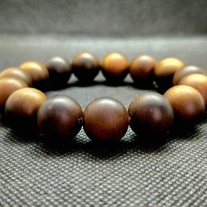 Könnte beinhalten: Ein Armband aus Holzperlen, kreisförmig angeordnet. Die Perlen sind in verschiedenen Brauntönen gehalten, von hellem Beige bis zu dunklem Braun. Das Armband besteht aus glatten, runden Perlen und ist zum Tragen am Handgelenk bestimmt.