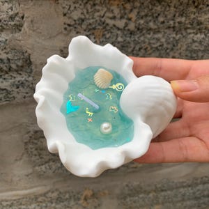 Könnte beinhalten: Weiße Keramik-Muschel-Schale mit einem blauen Harz-Innenraum, gefüllt mit Glitzer und Muscheln. Die Schale ist in Form einer Venusmuschel geformt und hat einen gewellten Rand.