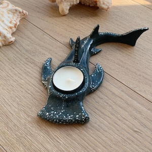 Puede incluir: Un portavelas de cerámica en forma de ballena de color verde azulado oscuro con una vela blanca en el centro. El portavelas tiene motas blancas y está sobre una superficie de madera. Conchas marinas y coral en el fondo.
