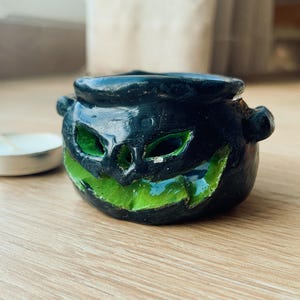 May include: A black ceramic Halloween cauldron with a carved jack-o'-lantern face, featuring green eyes and a grinning mouth. The cauldron has two small handles and sits on a wooden surface. A small tea light is in the background.
