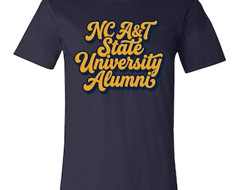 Könnte beinhalten: Ein kurzärmeliges T-Shirt in Marineblau mit dem gelben Schriftzug NC A&T State University Alumni in einer fetten, kursiven Schriftart, die mittig auf der Brust platziert ist. Die Schrift hat einen dunklen Schatten.