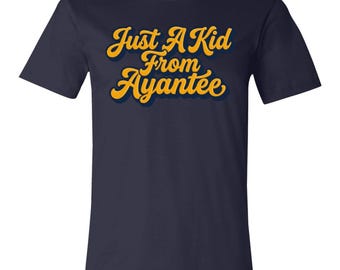 Könnte beinhalten: Ein kurzärmeliges T-Shirt in Marineblau mit dem Schriftzug Just A Kid From Ayantee in einer fetten, goldgelben Schreibschrift auf der Brust. Die Buchstaben haben einen dezenten dunkelblauen Schlagschatten auf dem Stoff.