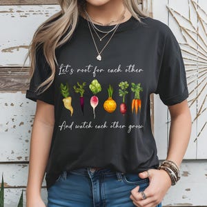 May include: Black t-shirt with the text "Let's root for each other And watch each other grow." The shirt features a watercolor illustration of various vegetables, including carrots, radishes, and onions.