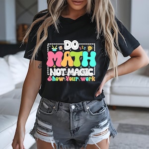 May include: Black t-shirt with the text "DO MATH NOT MAGIC Show Your Work" in colorful bubble letters. The shirt features a math-themed design with numbers, symbols, and a lightbulb. The person is wearing gray denim shorts.