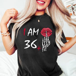 Könnte beinhalten: Schwarzes T-Shirt mit dem Text "I AM 36+". Das Design zeigt eine rote Glitzerlippe und eine Skeletthand, die den Mittelfinger zeigt. Die Zahl 36 ist in Weiß.