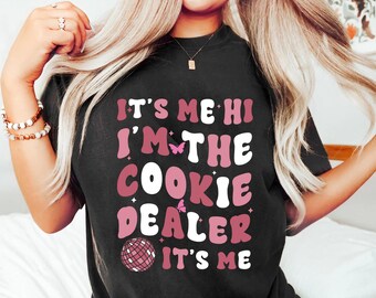 May include: Black t-shirt with the text "IT'S ME HI I'M THE COOKIE DEALER IT'S ME" in pink and white lettering. The design includes butterfly and disco ball graphics. The shirt is a casual, comfortable style.
