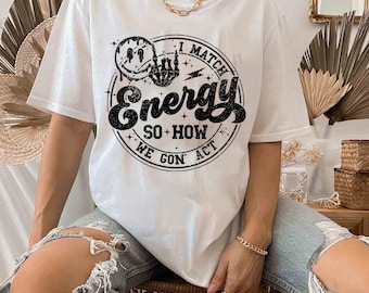 May include: White t-shirt with a black graphic that reads "I Match Energy So How We Gon Act". The design includes a smiley face, a hand with a raised middle finger, and lightning bolts. The shirt is worn with ripped blue jeans.